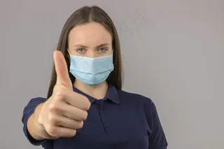 身穿蓝色马球衫的年轻女子，戴防护性医用面罩，站在浅灰色背景上，大拇指朝上，表情积极