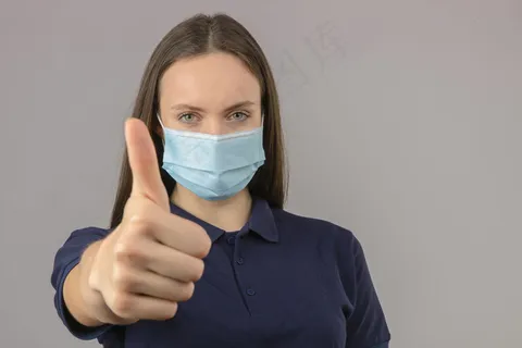 身穿蓝色马球衫的年轻女子,戴防护性医用面罩,站在浅灰色背景上,大拇指朝上,表情积极 身穿蓝色马球衫的年轻女子,戴防护性医用面罩,站在浅灰色背景上,大拇指朝上,表情积极
