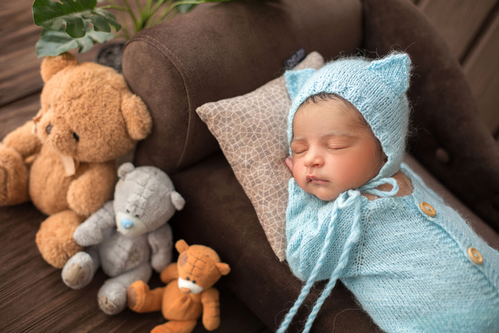 男婴新生婴儿穿着蓝色钩针睡衣睡在棕色沙发上，周围有三只玩具熊