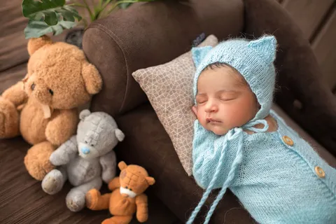 男婴新生婴儿穿着蓝色钩针睡衣睡在棕色沙发上,周围有三只玩具熊 男婴新生婴儿穿着蓝色钩针睡衣睡在棕色沙发上,周围有三只玩具熊