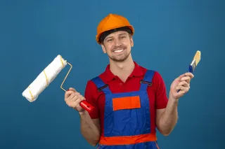 穿着建筑制服，戴着安全帽的年轻建筑工人拿着油漆辊和刷子，脸上挂着灿烂的笑容