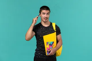 身穿深色t恤的男生正面图黄色背包拿着文案笔和文件在浅蓝色墙上思考
