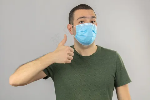 戴着医用面罩的年轻人做着ok的手势,拇指向上,手指分开放在白色上 戴着医用面罩的年轻人做着ok的手势,拇指向上,手指分开放在白色上