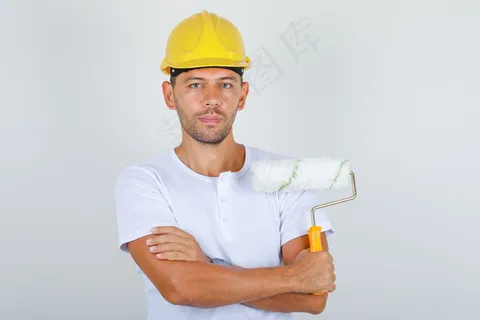 手拿油漆辊的建筑工人交叉双臂穿着白色t恤,头盔,正面视图。 手拿油漆辊的建筑工人交叉双臂穿着白色t恤,头盔,正面视图。