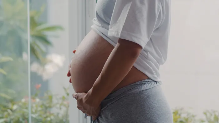年轻的亚洲孕妇抱着肚子和孩子说话。妈妈开心的笑着积极平和地照顾宝宝，怀孕时在家客厅靠窗的地方。