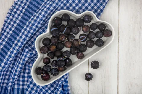 在灰色木质背景上，蓝色格子桌布上的白色碗上的酸蓝色黑色小树莓俯视图