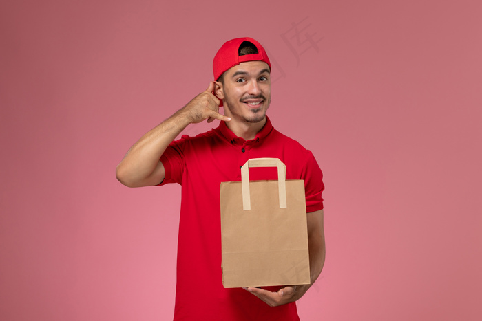 正面图：身穿红色制服披肩的年轻男性信使手持粉色背景上的纸质食品包。