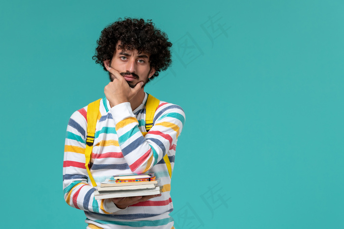 身穿条纹衬衫、背着黄色背包、拿着书、在蓝色墙上思考的男学生正面图