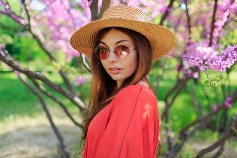清新的春天写真可爱的微笑女人穿着时髦的珊瑚色连衣裙，戴着草帽享受阳光