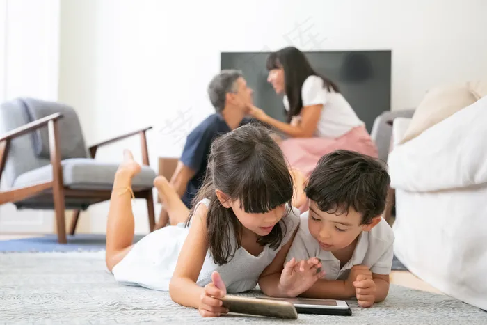 专注的小孩躺在客厅的地板上，在父母接吻时使用带有学习应用程序的数字设备