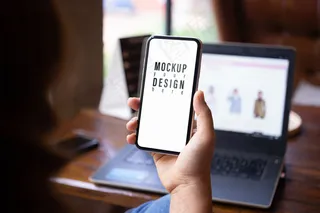 Mokcup手机。一个人拿着智能手机和模糊笔记本电脑放在咖啡馆的木桌上。