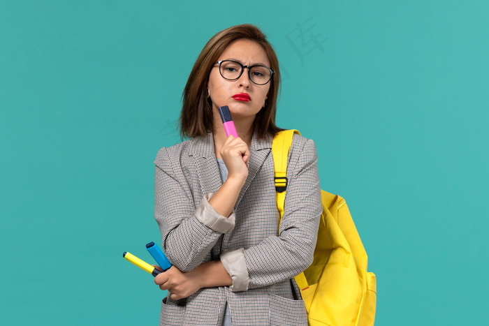身穿灰色夹克、背着黄色背包、拿着毛毡笔的女学生正对着淡蓝色的墙上思考