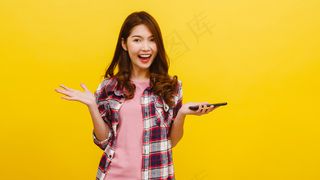 照片中的亚洲女性惊讶地使用手机，表情积极，穿着休闲服，看着黄色墙上的相机。快乐可爱快乐的女人为成功而高兴。