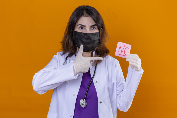 中年女性医生身穿黑色防护面罩，用听诊器拿着提醒纸，手指上没有手指指着孤立的橙色背景。