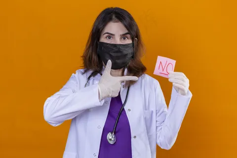 中年女性医生身穿黑色防护面罩，用听诊器拿着提醒纸，手指上没有手指指着孤立的橙色背景。