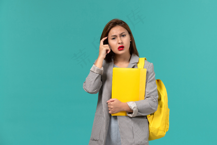 身穿灰色夹克、背着黄色背包、拿着文件的女学生在淡蓝色墙上思考的正面图
