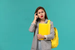 身穿灰色夹克、背着黄色背包、拿着文件的女学生在淡蓝色墙上思考的正面图