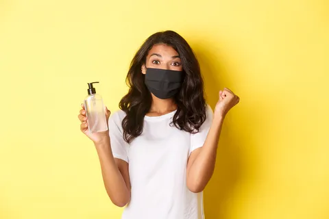 covid-19 的概念、社交距离和生活方式。戴着面罩的快乐非洲裔美国女性的形象,对创立好的洗手液感到高兴,在黄色背景下欢欣鼓舞 covid-19 的概念、社交距离和生活方式。戴着面罩的快乐非洲裔美国女性的形象,对创立好的洗手液感到高兴,在黄色背景下欢欣鼓舞