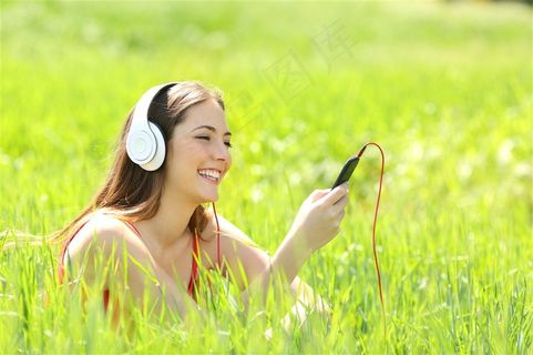 草地里戴白色耳机看手机的外国女性高清摄影图片